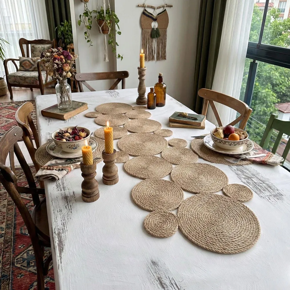 Asymmetric jute table runner of coiled discs on a white cloth dining table, with candles, stoneware plates and a woven wall hanging.