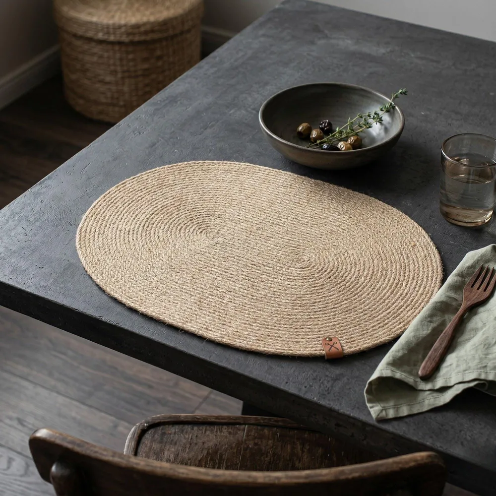 Handwoven oval jute placemat shown in full on a dark wood table with a green bowl, natural braided weave.