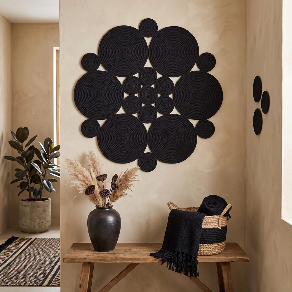 May include: A large wall art piece composed of multiple black circular rope coils arranged in a floral pattern. The design features varying sizes of concentric circles, creating a textured, geometric display mounted on a light beige wall.