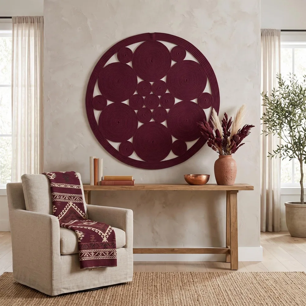 Large burgundy cotton rope wall mandala on a plaster wall above a cream armchair, with a patterned throw and dried stems.
