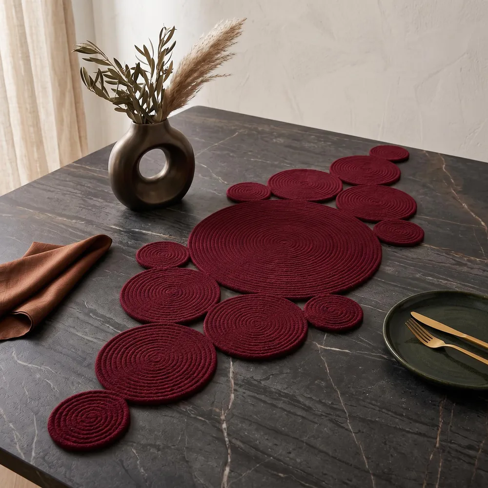 Burgundy cotton rope table runner made of graduated coiled discs, laid on a dark wood table with a Christmas tree, warm string lights, a burgundy candle, dried pampas grass, and orange slices in the background.